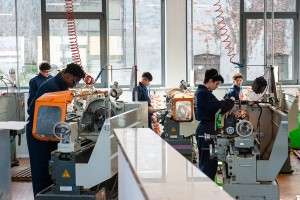 Studenti in tuta da lavoro blu utilizzano torni e fresatrici in un'officina meccanica ben illuminata, impegnati in attività di lavorazione del metallo.