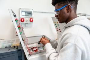 Un giovane studente indossa occhiali protettivi mentre prepara un filo con delle forbici per lavorare su un quadro elettrico sullo sfondo, con vari cavi colorati e componenti elettronici all'interno.
