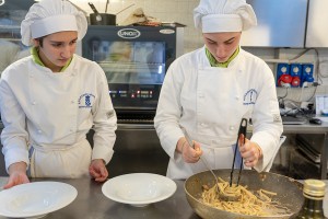 Due studentesse vestite con tenuta da chef che impiattano della pasta appena preparata all'interno del laboratorio di cucina