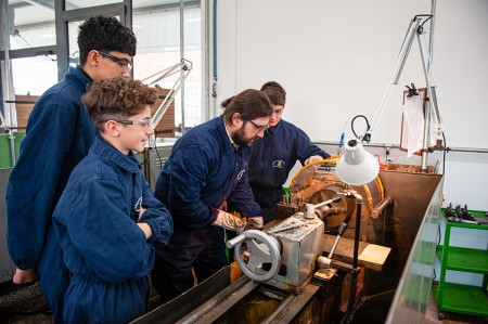 Gruppo di studenti in tuta da lavoro blu mentre osservano un insegnante che opera su una macchina utensile in un laboratorio tecnico.