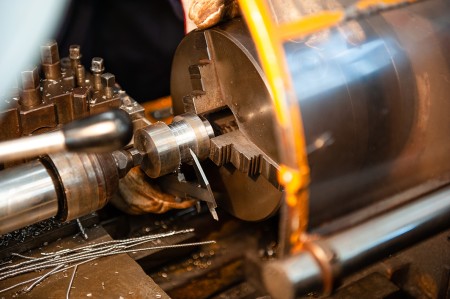 Primo piano di una mano guantata che misura tramite calibro un pezzo di metallo montato su un tornio, mentre vengono eseguite operazioni di precisione di tornitura.