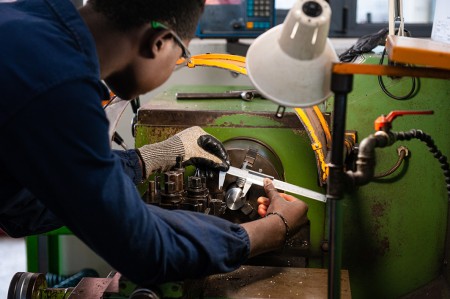 Primo piano di un ragazzo che misura tramite calibro un pezzo di metallo montato su un tornio, mentre vengono eseguite operazioni di precisione di tornitura.
