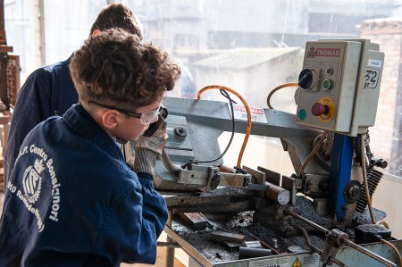 Studente in tuta da lavoro blu utilizza una macchina utensile in un laboratorio meccanico, operando con attenzione sulla manovella della macchina.