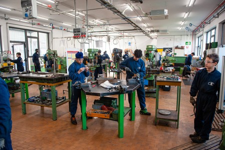 Studenti in tuta da lavoro blu utilizzano torni e fresatrici in un'officina meccanica ben illuminata, impegnati in attività di lavorazione del metallo.