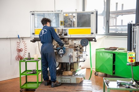 Studente in tuta da lavoro blu utilizza una macchina utensile in un laboratorio meccanico, operando con attenzione sulla manovella della macchina.