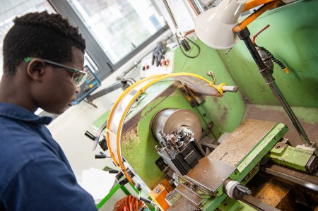 Studente in tuta da lavoro blu utilizza una macchina utensile in un laboratorio meccanico, operando con attenzione sulla manovella della macchina.