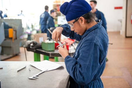 Studente in tuta da lavoro blu e con occhiali protettivi misura tramite il calibro un pezzo di metallo nel laboratorio meccanico
