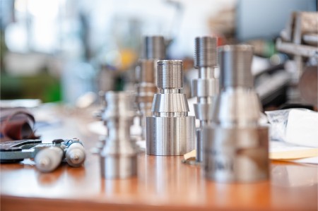 Primo piano di componenti meccanici lavorati in metallo, con filettature e forme precise, disposti su un tavolo in un ambiente di officina.