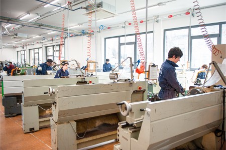 Un gruppo di studenti in tuta blu lavora sui torni in un'officina meccanica spaziosa e ben attrezzata, con postazioni individuali illuminate da grandi finestre.