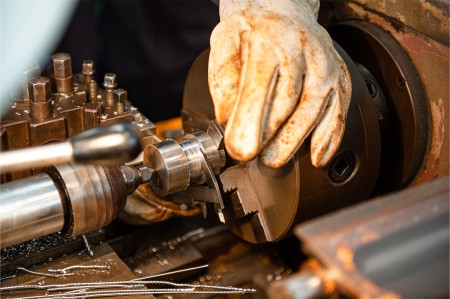 Primo piano di una mano guantata che lavora su un pezzo di metallo montato su un tornio, mentre vengono eseguite operazioni di precisione di tornitura.