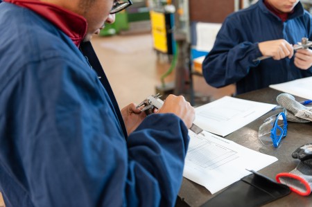Studente in tuta da lavoro che utilizza un calibro per misurare un componente meccanico, con fogli tecnici sul banco e occhiali di protezione a fianco.