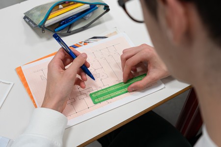 Uno studente seduto al tavolo disegna un impianto elettrico su carta millimetrata mediante matita squadre e normografo