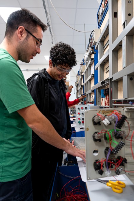 Insegnante assiste uno studente durante una lezione pratica di impianti elettrici industriali, spiegando come collegare correttamente i cablaggi.