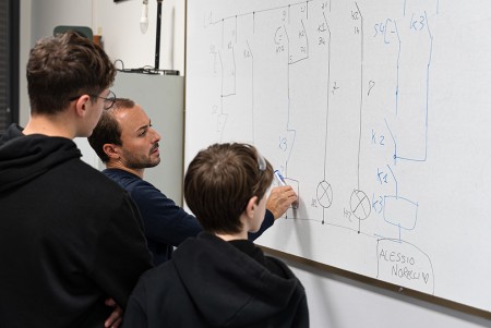 Insegnante indica un diagramma alla lavagna durante una lezione di cablaggio elettrico, spiegando i dettagli a due studenti.