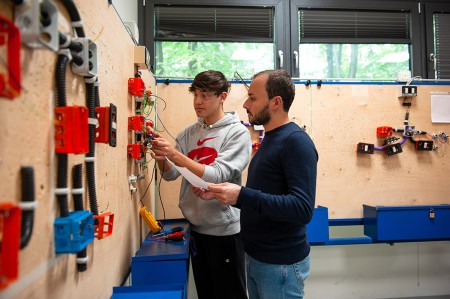 Insegnante assiste uno studente durante una lezione pratica di cablaggio elettrico, spiegando come collegare correttamente i fili in una scatola di derivazione.