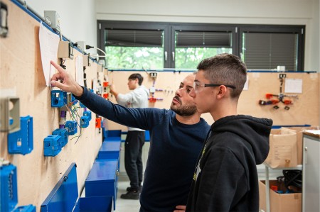Insegnante spiega a uno studente un circuito elettrico su una parete didattica, durante una lezione pratica in laboratorio.