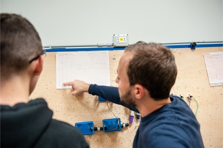 Insegnante indica un diagramma su carta durante una lezione di cablaggio elettrico, spiegando i dettagli a uno studente.