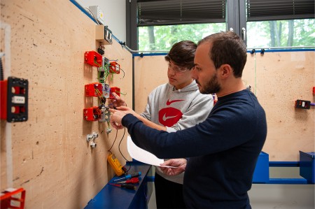Insegnante assiste uno studente durante una lezione pratica di cablaggio elettrico, spiegando come collegare correttamente i fili in una scatola di derivazione.