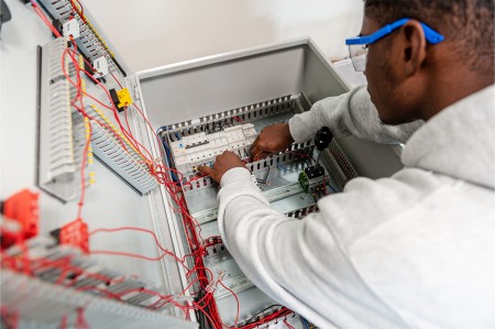Un giovane studente indossa occhiali protettivi mentre lavora su un quadro elettrico aperto, con vari cavi colorati e componenti elettronici all'interno. Il ragazzo sta effettuando collegamenti o verifiche su un pannello di controllo elettrico.