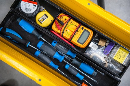 Una cassetta degli attrezzi contenente vari strumenti, tra cui cacciaviti, metro a nastro, un multimetro digitale e del nastro isolante.
