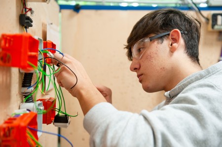Un ragazzo con occhiali protettivi sta installando i cablaggi elettrici all'interno di un impianto civile, fissando i tubi corrugati e organizzando i fili all'interno di una scatola di derivazione.