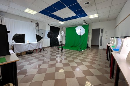 Studio fotografico scolastico con attrezzatura professionale: schermi bianchi e verdi, softbox e ombrelli per l'illuminazione, e vari box per la fotografia di prodotto su un tavolo laterale.
