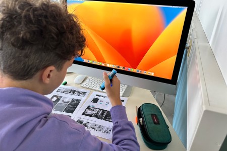 Studente al lavoro su un progetto grafico, con fogli stampati e un evidenziatore, seduto davanti a un computer con schermo acceso su interfaccia Mac.