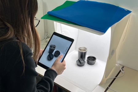 Studentessa fotografa con un tablet degli oggetti esposti in un box fotografico illuminato, con uno sfondo bianco e accessori posizionati per la fotografia di prodotto.