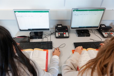 Due studenti lavorano su esercizi di contabilità con computer e calcolatrici, consultando libri di testo in un'aula.