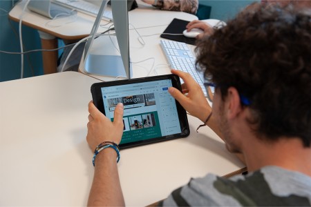 Uno studente lavora su un progetto di web design utilizzando un tablet, mentre altri studenti usano computer in un'aula attrezzata.