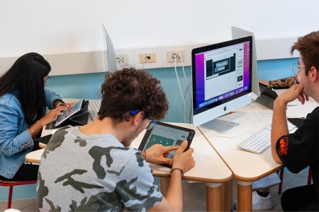 Tre studenti lavorano a un progetto di design, utilizzando tablet e computer desktop in un'aula attrezzata con postazioni informatiche.